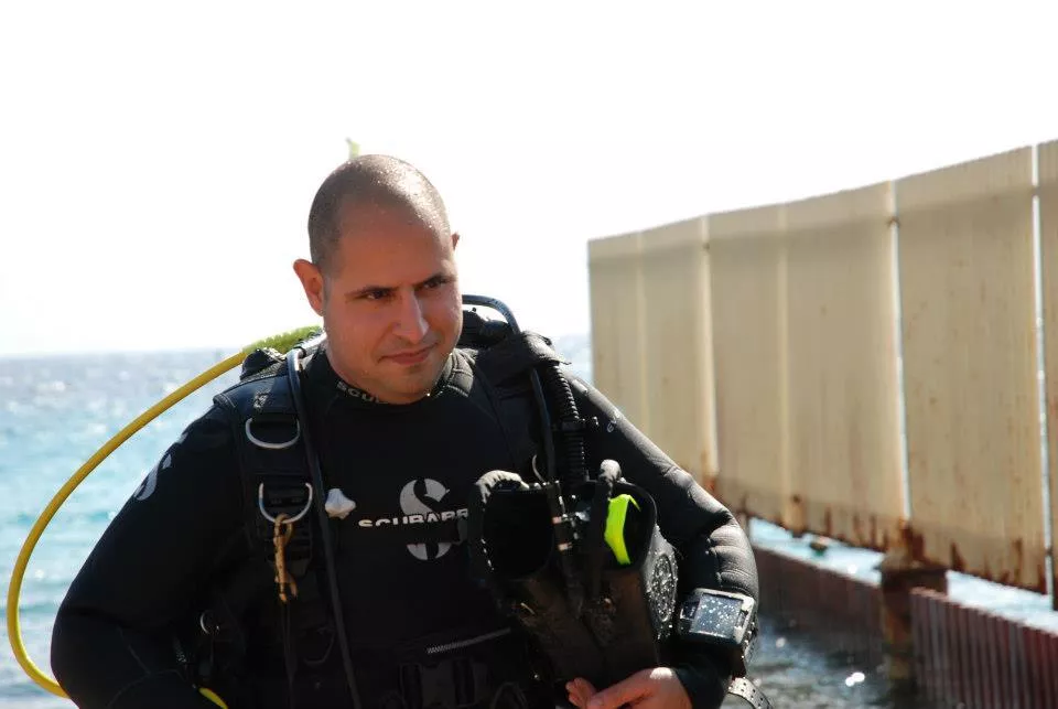 קובי חי יוצא מצלילה, ברקע חוף אלמוג וגדר שמבודדת את שמורת האלמוגים באילת