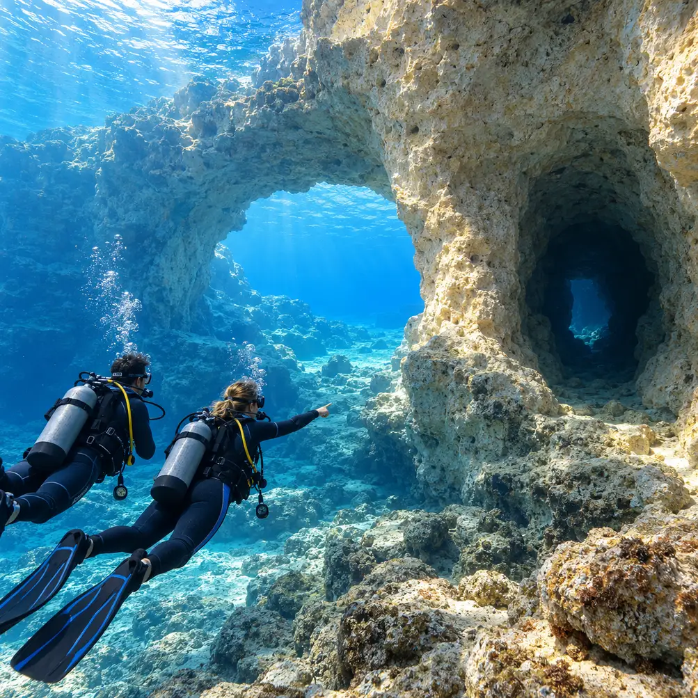 Two scuba divers examine an open rock arch beside a narrower, darker passage before choosing their route.