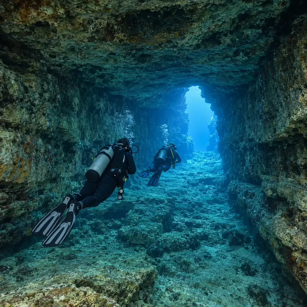 Two scuba divers swim through a broad rock tunnel toward a bright opening into open water.